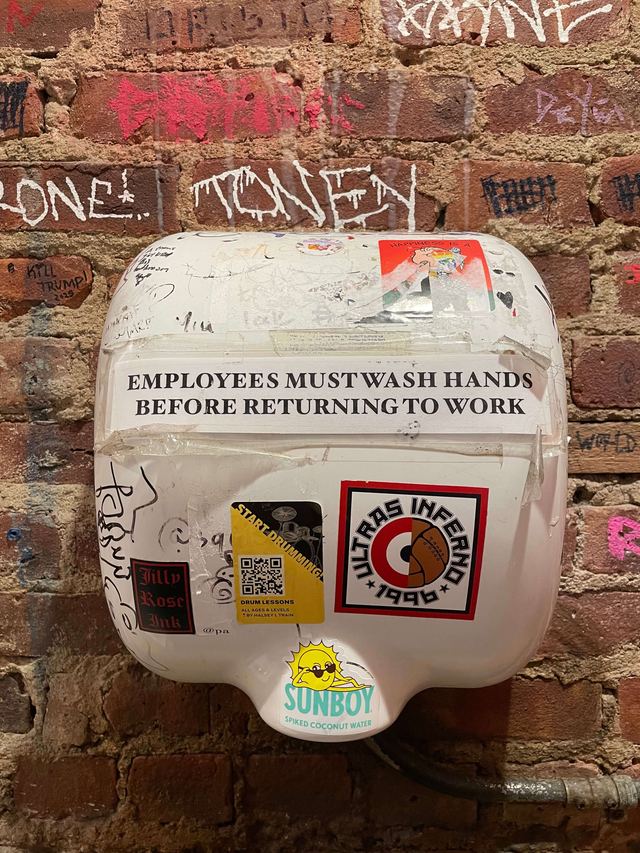 Sign printed on a sticker-covered hand dryer against a graffitied brick wall at Father Knows Best