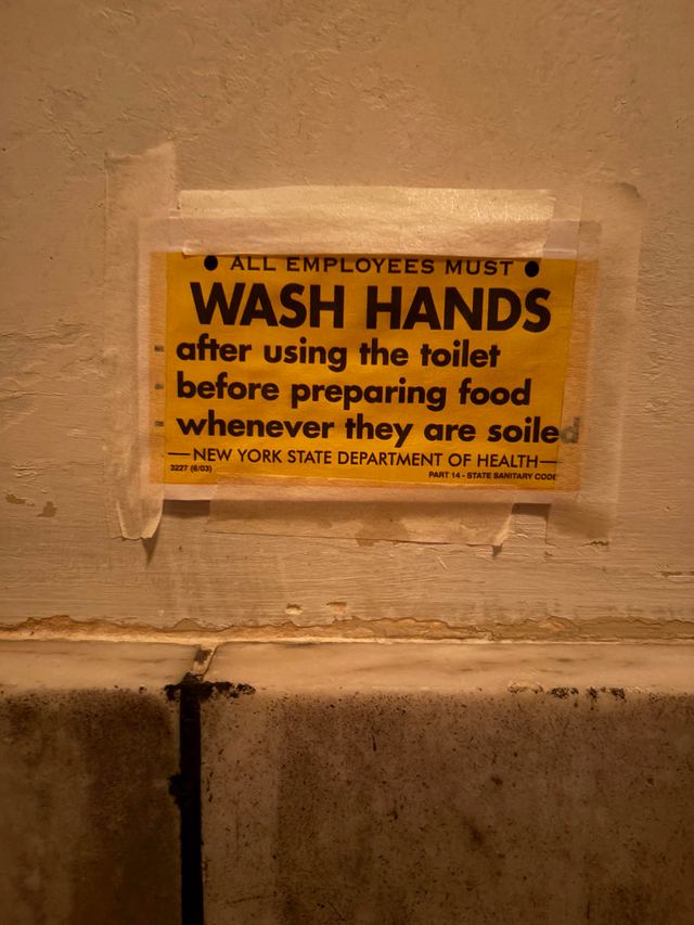 Yellow New York State Department of Health placard taped to a painted wall, listing when employees must wash hands