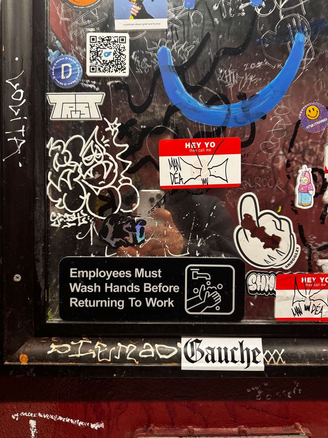 Engraved black sign on a Brooklyn bathroom door covered in graffiti, stickers and a painted blue smile