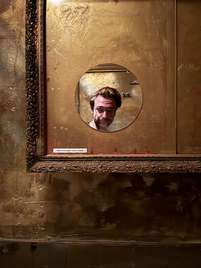 Small sign beneath a round mirror in an ornate gold frame, reflecting the photographer, in Brooklyn