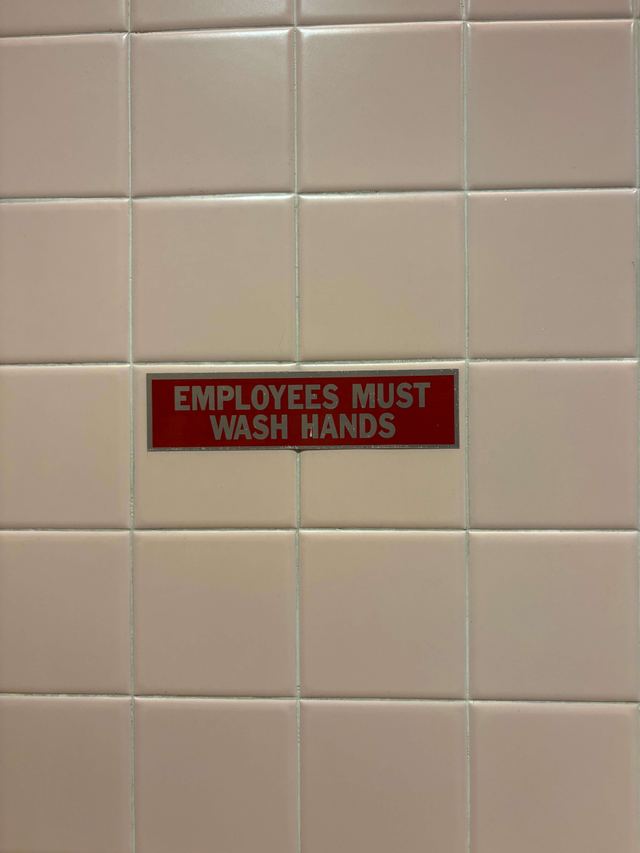 Small red and silver “Employees Must Wash Hands” plate on cream tile at a Broadway theater